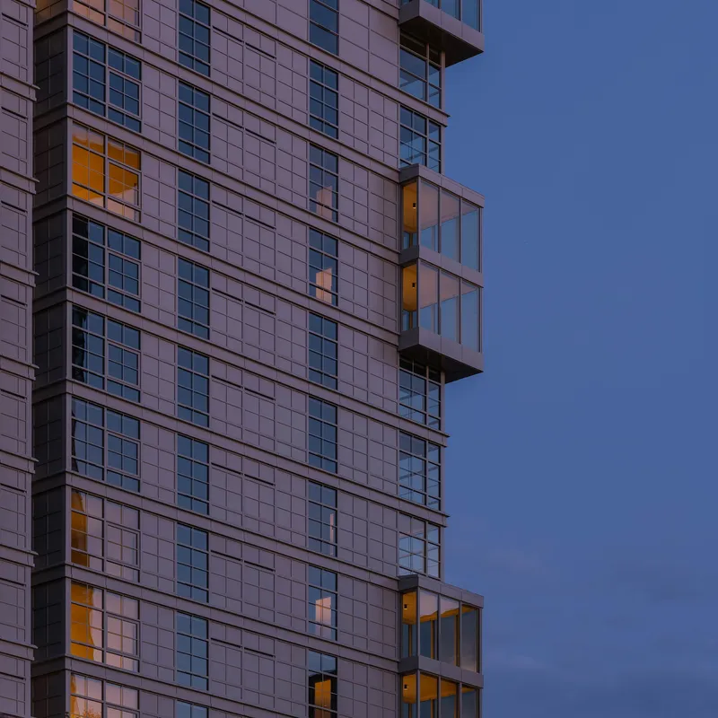 Gridded facade with projecting glass balconies illuminated at dusk against a deepening blue sky