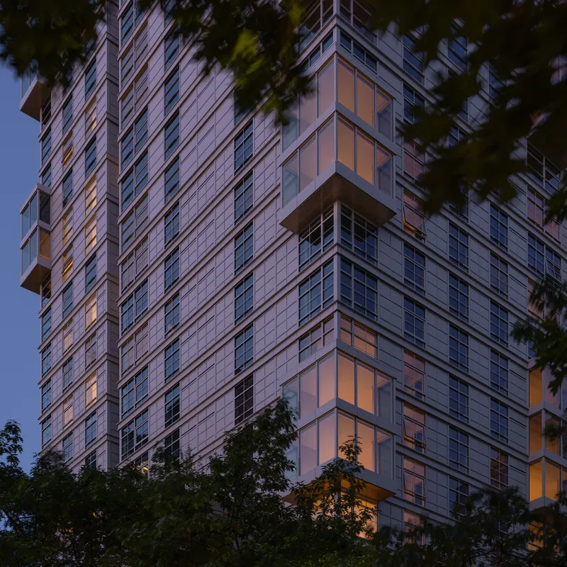 Glass and panel facade of a residential tower framed by trees at dusk with interior lights glowing