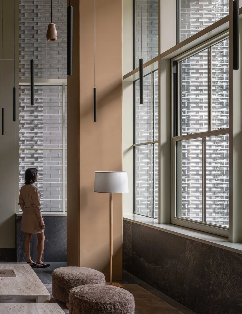 Double-height lobby with tall windows framed by textured brick walls as a person stands in afternoon light