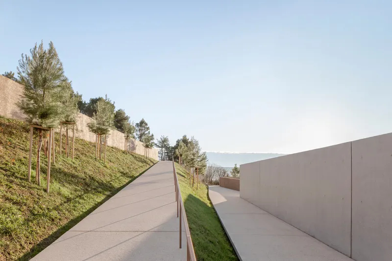 Concrete ramp ascending hillside between planted slope and smooth retaining wall with distant lake