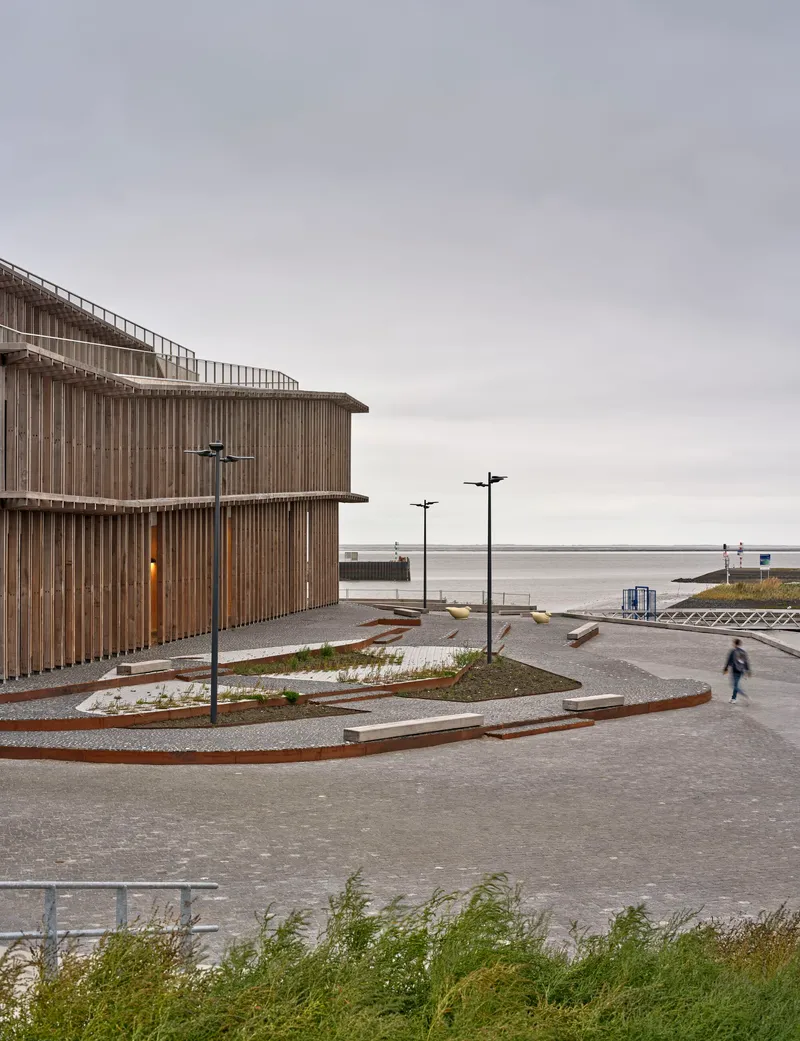 View of the curved timber facade with terraced paving and a lone figure walking along the waterfront