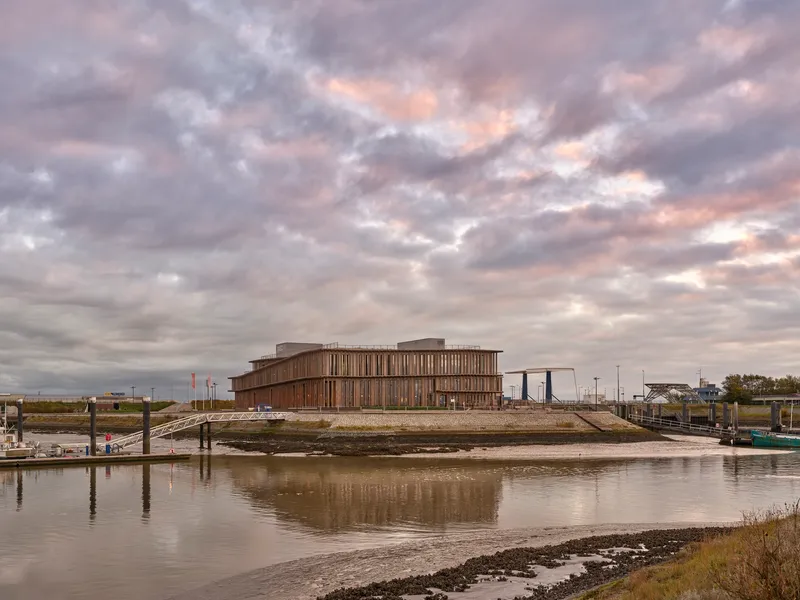 Horizontal timber-clad building reflected in the tidal waters of a mudflat harbor under dramatic clouds