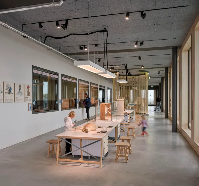 Exhibition hall with concrete plank ceiling, work tables, wooden stools, and children engaging with displays