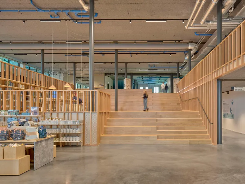 Wide timber staircase ascending past a retail display wall under an exposed concrete soffit with mechanical ducts