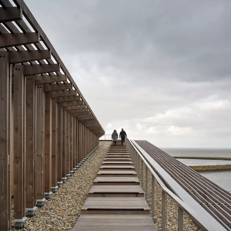 Timber boardwalk with open stairway alongside vertical slat screen and gravel ballast roof overlooking the sea