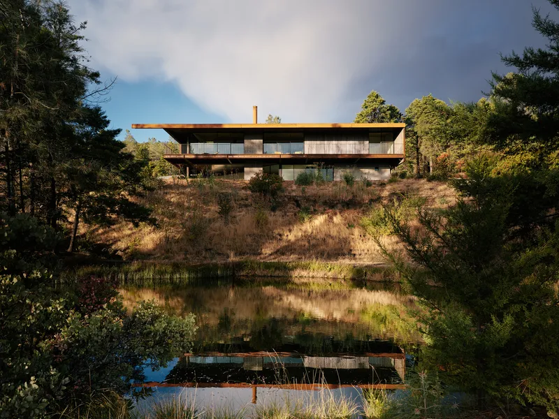 Two-story house with flat roof reflected in a still pond at dusk beneath dramatic cloudy skies