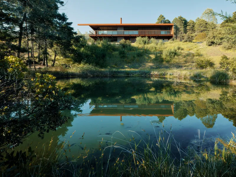 Two-storey horizontal volume with deep overhangs reflected in a natural pond surrounded by grasses and pines