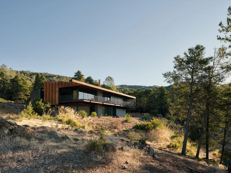 Sloped metal roof over cantilevered volume on a rocky hillside with sparse vegetation and distant ridges