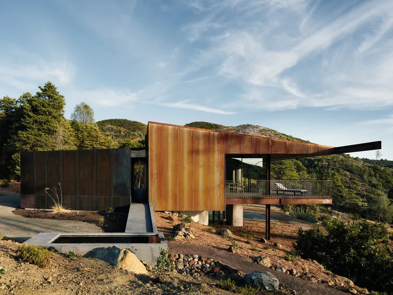 Corrugated metal facade with vertical fins next to weathered steel panels on pilotis above exposed bedrock