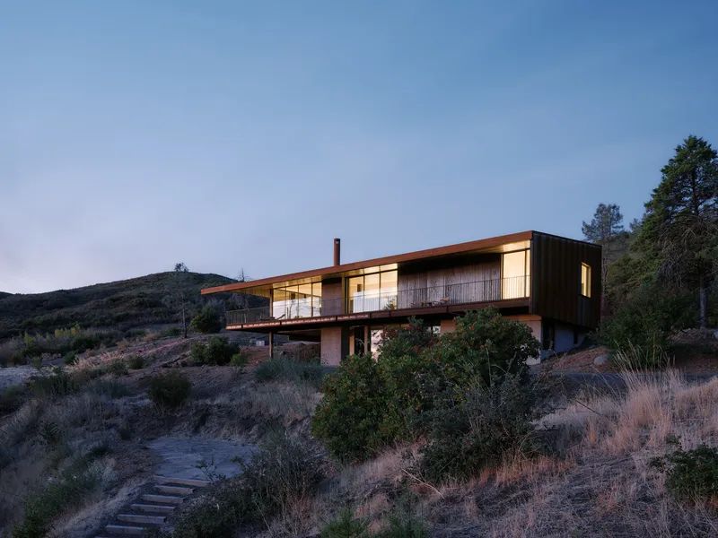 Two-level residence with corten upper volume and timber lower level nestled in scrubland at twilight