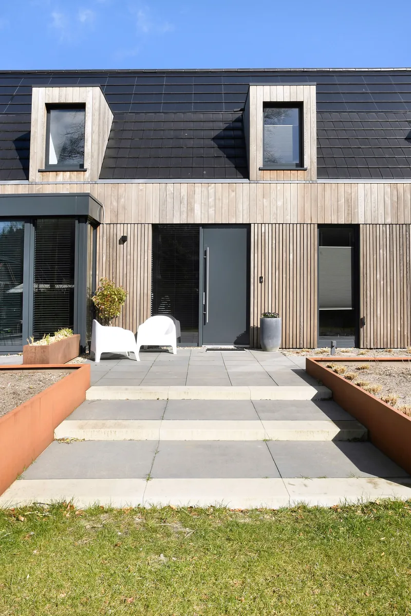 Entrance facade with vertical timber cladding, black tile roof and concrete stepping stones in midday sun