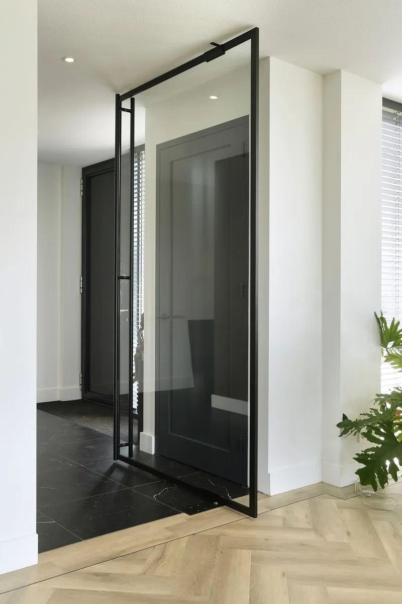 Pivoting glass door with black steel frame opening from herringbone oak floor to dark tile entry