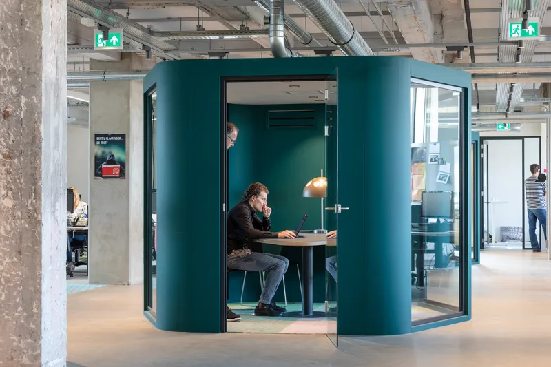 Freestanding teal-painted meeting pod with glass doors in an open office with exposed services