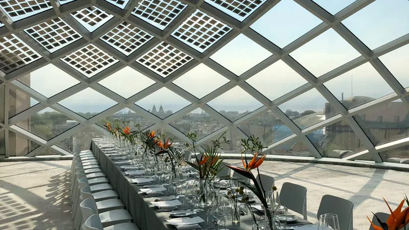Interior dining hall beneath a diamond-patterned glazed roof structure overlooking the city in hazy daylight