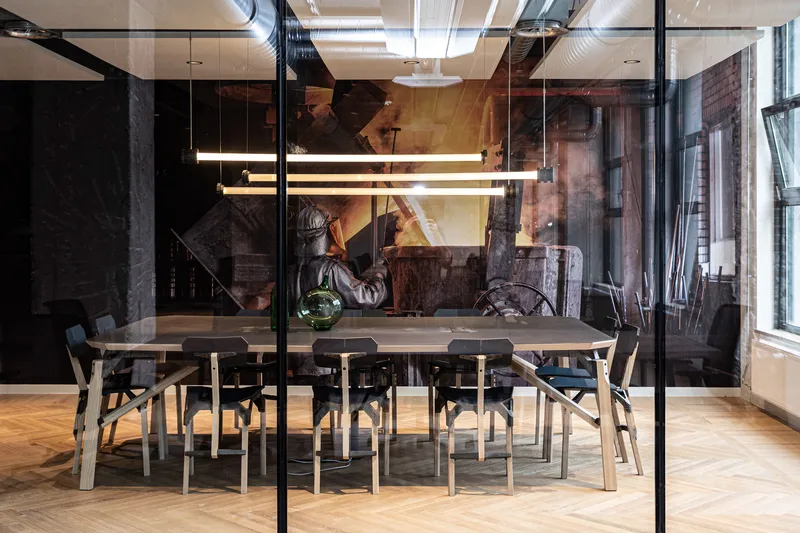 Glass-partitioned meeting room with timber furniture and parquet flooring beneath exposed ductwork and linear pendant lights