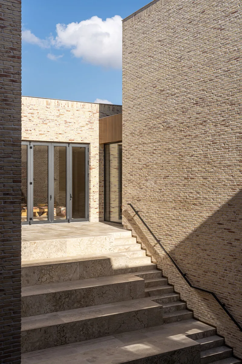 Stone staircase ascending between buff brick walls toward glazed doors beneath blue sky