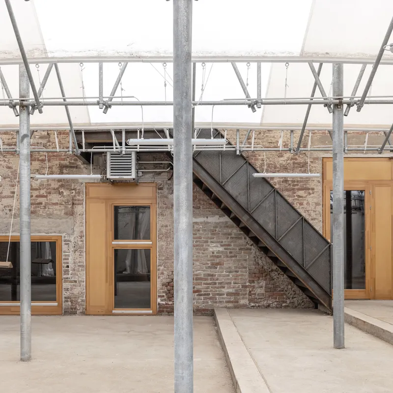 Exposed brick wall with timber-framed windows and dark metal staircase beneath galvanized steel roof trusses
