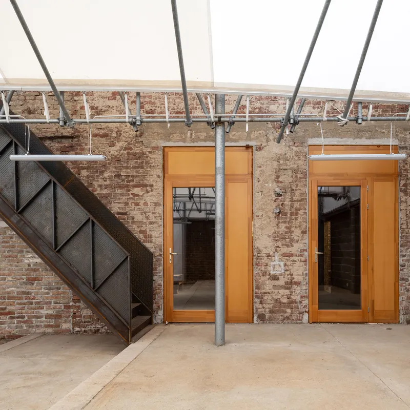 Two timber-framed glass doors set into weathered brick wall beneath exposed steel structure and translucent roof panels