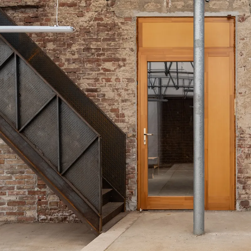 Orange timber door with glazed upper panel set within exposed brick wall beside a dark metal staircase