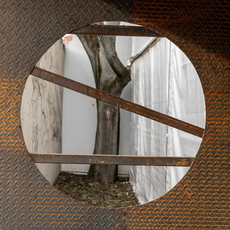 Circular opening in weathered steel diamond plate wall framing a tree and white fabric screen