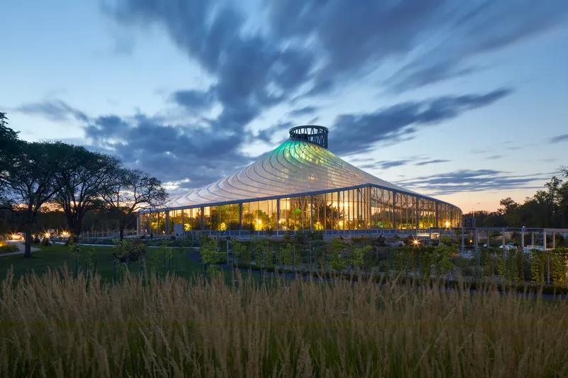 The illuminated glass pavilion with its conical roof at dusk beyond a field of dry grasses