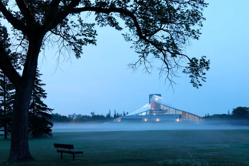 Distant view of the lit conservatory across a misty lawn with a bench and oak tree at dawn