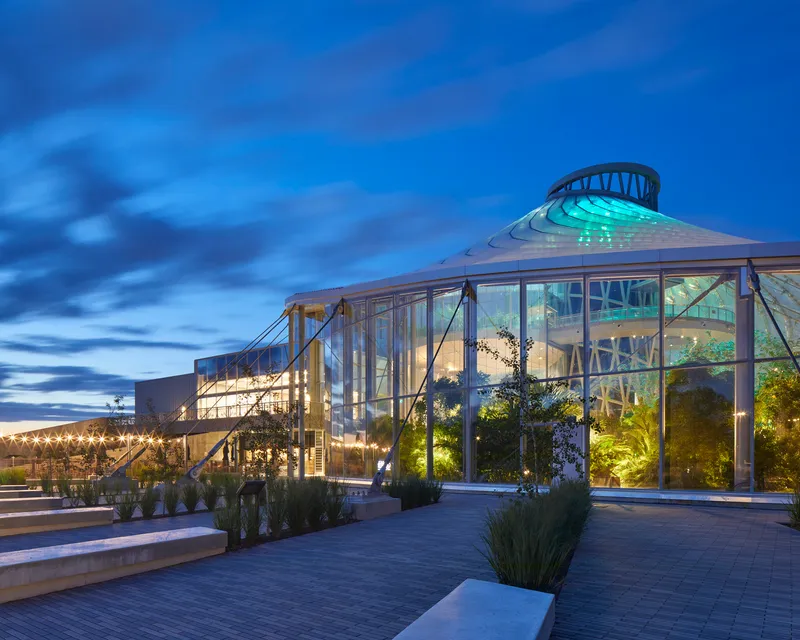 Glass conservatory volume with exposed steel structure and central dome illuminated against a blue evening sky
