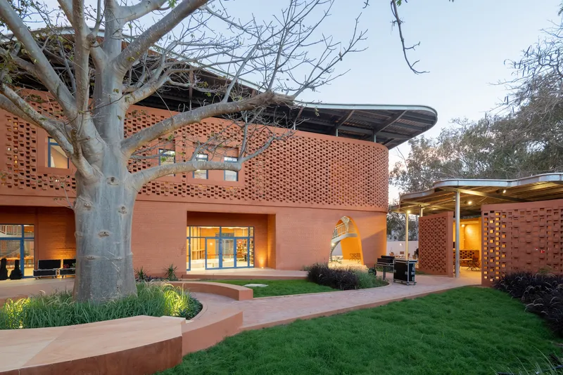 Perforated terracotta facade with curved volumes surrounding a courtyard garden with mature baobab tree at dusk