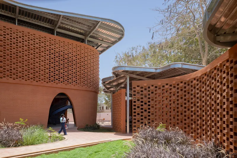 Arched passageway between terracotta volumes with undulating metal roof and planted beds under clear sky