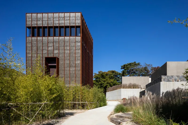 Timber-clad tower rising above a curved pathway bordered by bamboo and ornamental grasses under clear blue sky