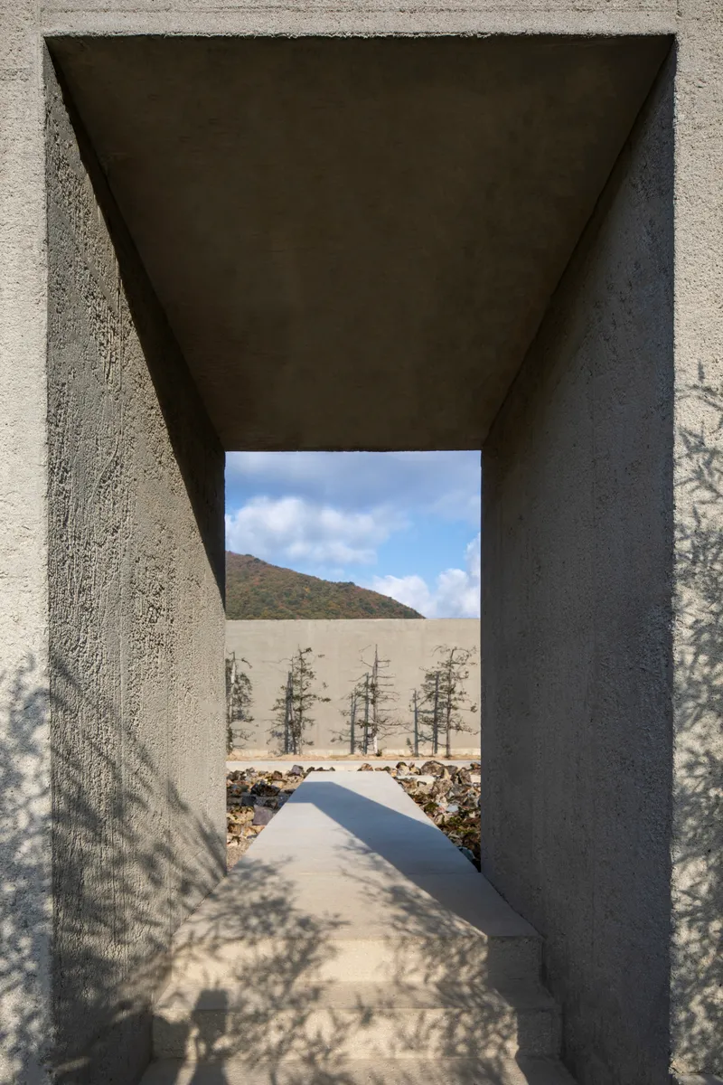 Tapered concrete portal framing a pathway toward young trees and distant hills under a cloudy sky