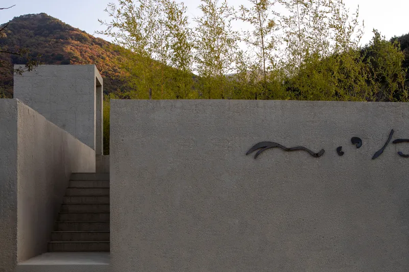 Textured concrete wall with calligraphic metal signage beside an ascending stair at dusk with trees beyond