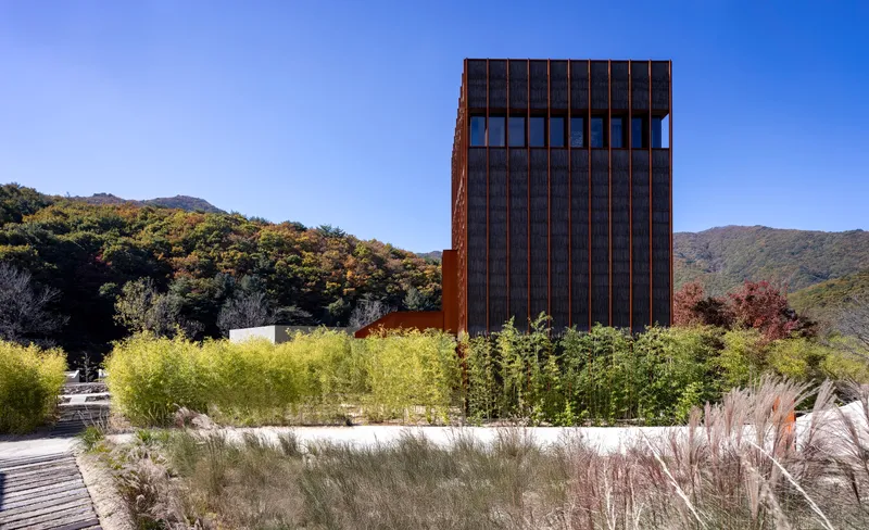 Corten steel tower rising above native grasses and planted beds with forested hills in the distance
