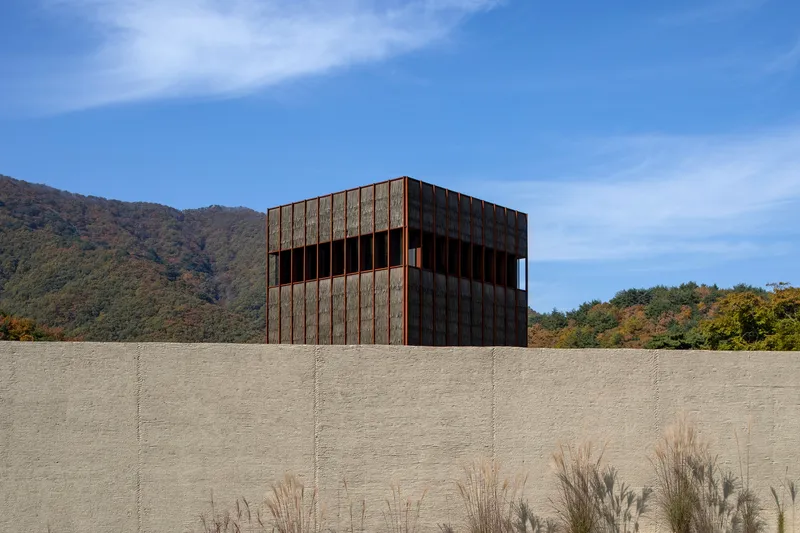 Weathered steel-clad volume above a textured concrete retaining wall with dry grasses in the foreground