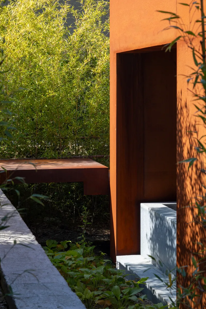 Rusted steel doorway framing bamboo and planted beds with afternoon shadows on the stucco wall