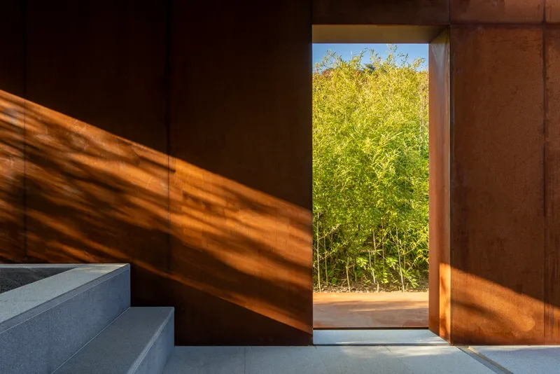 Interior view through rusted steel walls toward a framed opening revealing bamboo grove in daylight