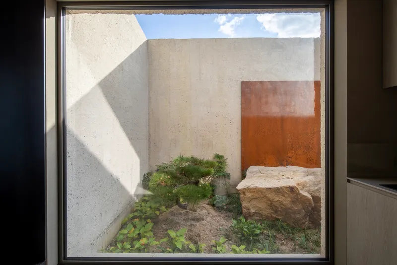 Framed courtyard view showing concrete walls, weathered steel panel, limestone boulder and planted greenery under blue sky
