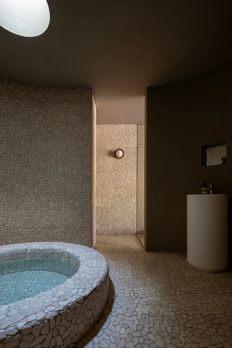 Bathing room with circular stone-clad soaking tub, pebble mosaic walls and cylindrical white basin under skylight