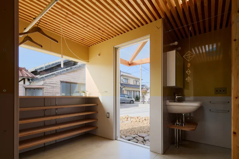 Entry corner with timber shelving and wall-mounted sink opening to a gravel courtyard and neighboring houses