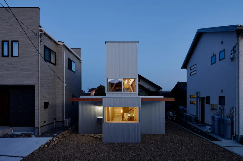 Corrugated metal clad volume with illuminated windows at dusk nestled between neighboring houses in gravel yard