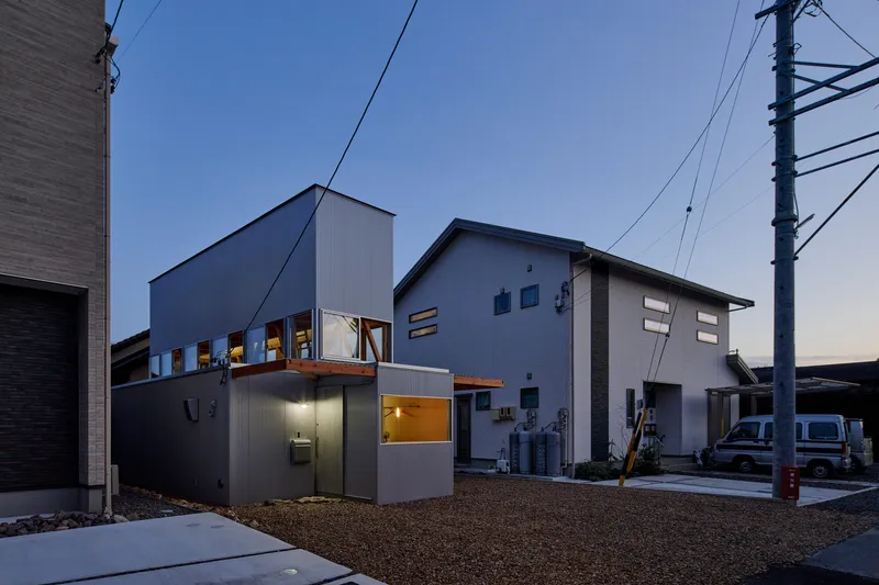 Street view showing the corrugated metal facade and upper glazed openings at twilight among adjacent residential buildings