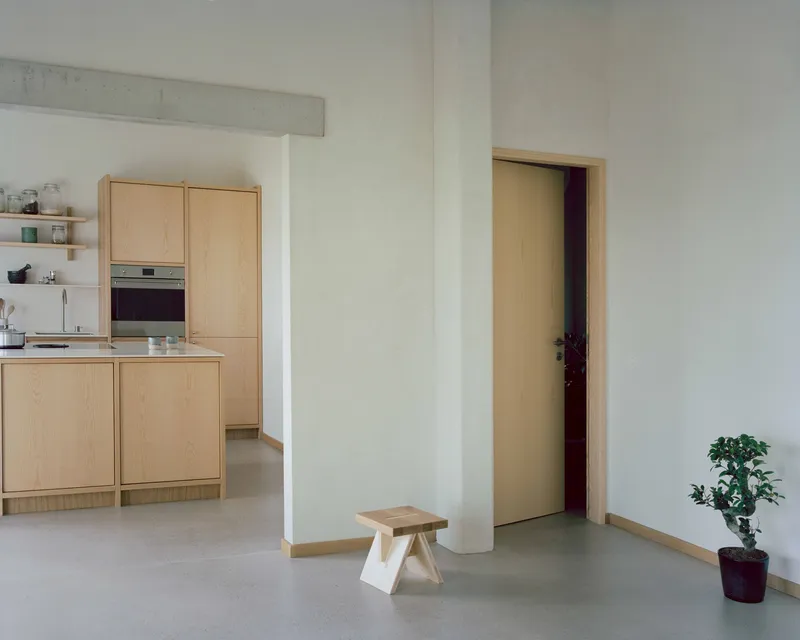 White-walled corridor with timber-framed doorway and small wooden stool beside a potted plant