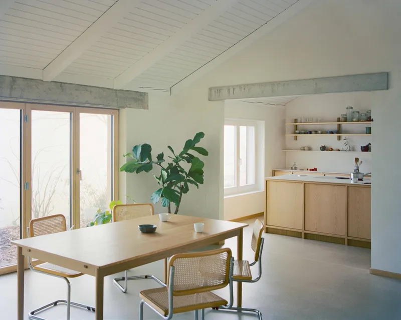 Dining area with exposed concrete lintel, timber cabinetry, and potted plants beside glazed doors