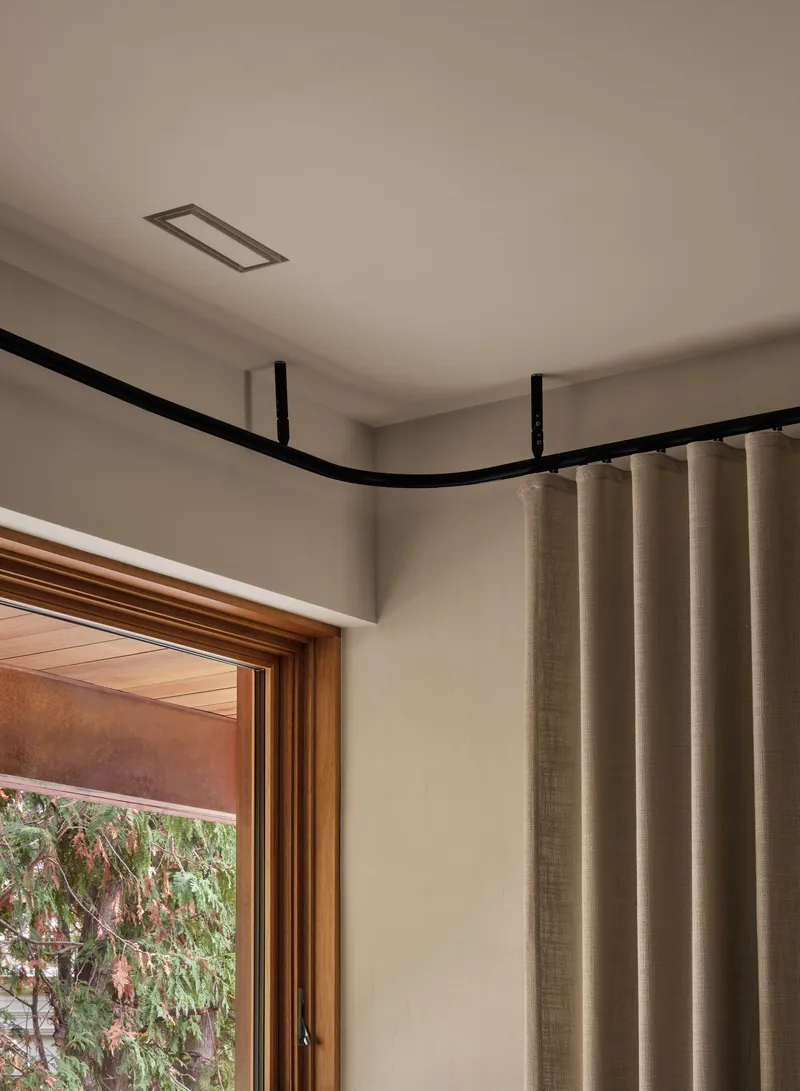 Corner detail of a curved black curtain track above a timber-framed window with garden view