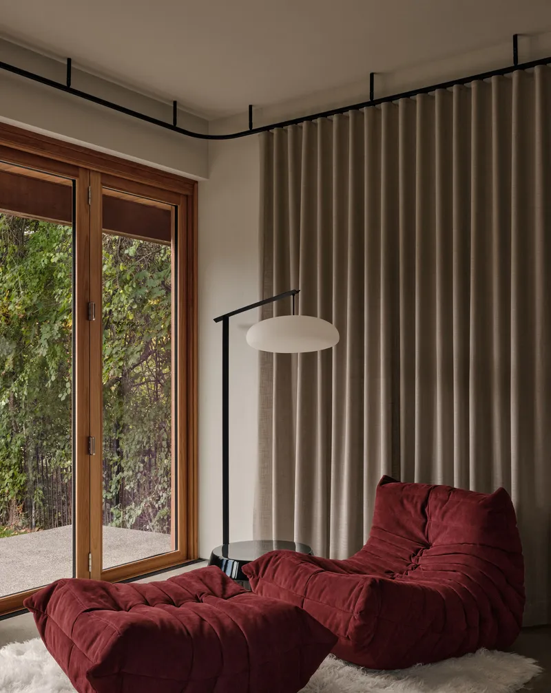 Reading nook with timber-framed glazing, beige curtain track and burgundy upholstered seating overlooking greenery