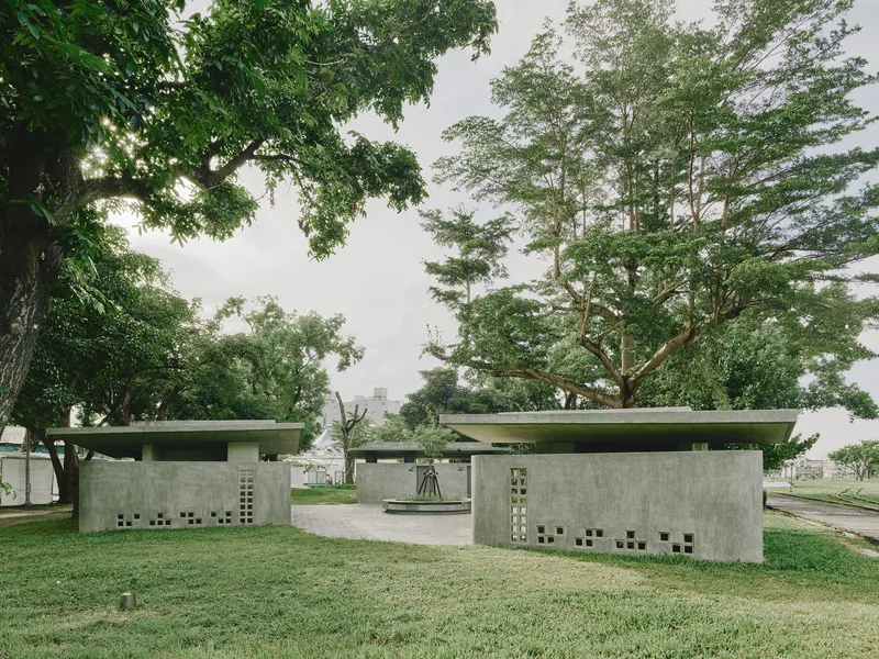 Pair of concrete pavilions with flat roofs and perforated walls beneath mature trees in a park