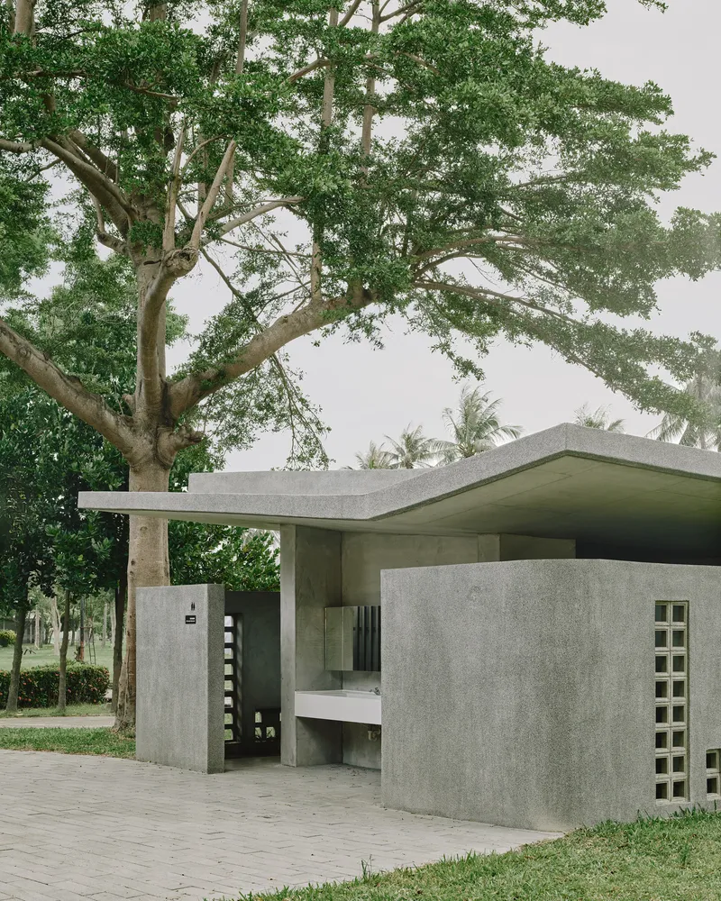 Corner view of concrete pavilion with cantilevered roof plane sheltering a bench beneath tree canopy