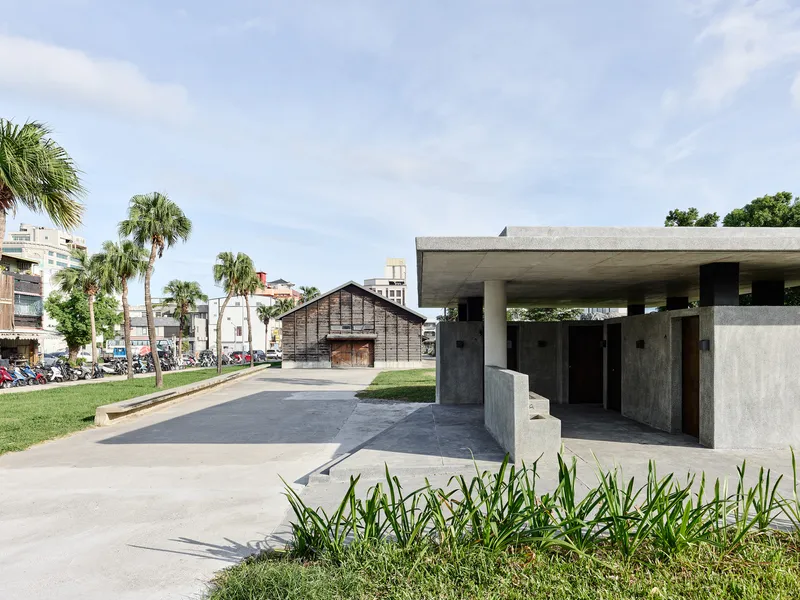 Concrete pavilion with flat roof and columned portico beside a lawn with palm trees and a timber structure