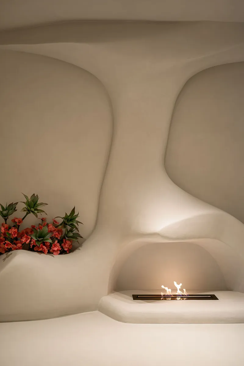 Curved plaster alcove with built-in fireplace and planted bed of flowering shrubs beneath warm ambient lighting