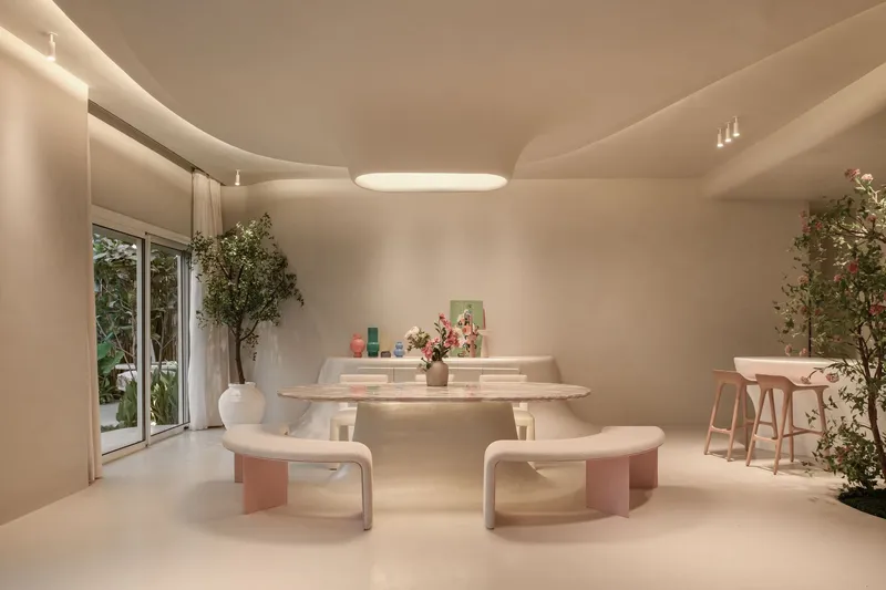 Dining area with a pale stone table, curved benches, and potted trees under soft ceiling light